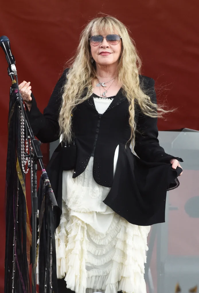 Picture of Stevie Knicks during a performance. Her is in her signature blonde waves and she wears a flowing black cardigan over a long and ruffled white dress. Around her neck are several moon necklaces in varying sizes. She also wears a pair of amber sunglasses. Even her mic is dressed up in Whimsigoth fashion, covered in clear beads and jewel-toned fabric.