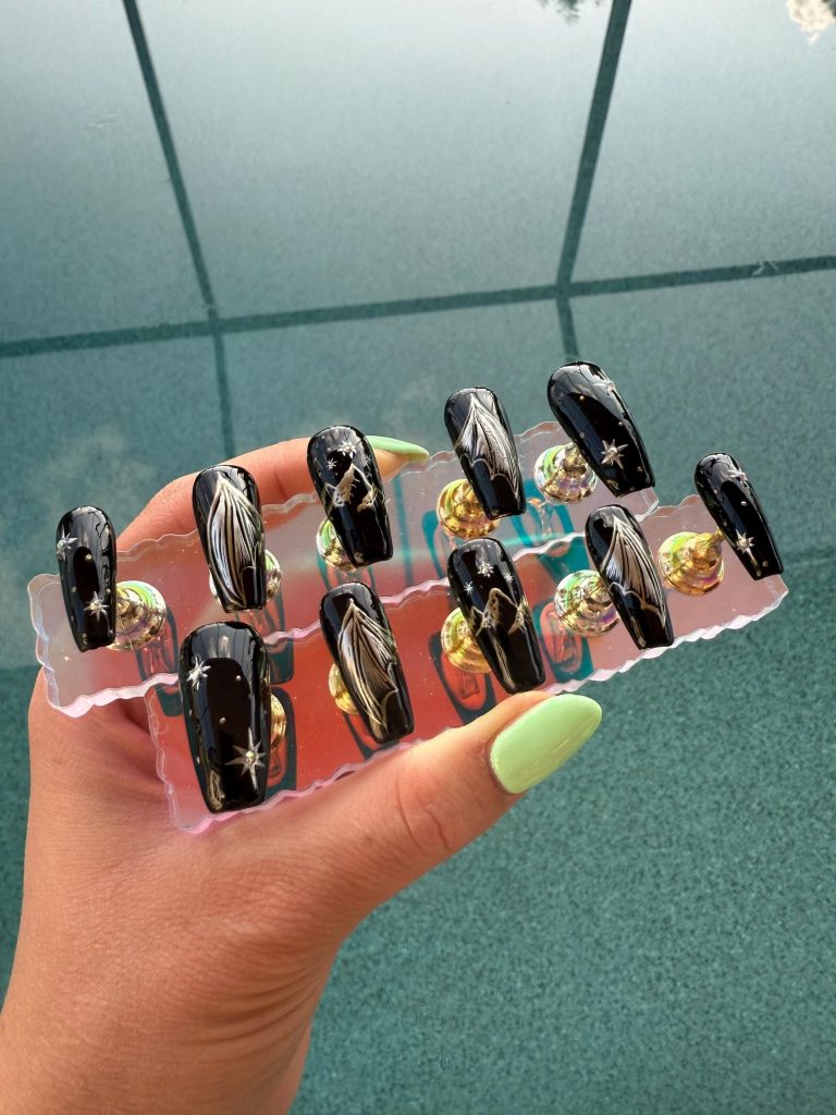 A set of press-on nails being held up by someone's hand. The press-on nails are black with golden accents featuring stars, mountains, and wings.