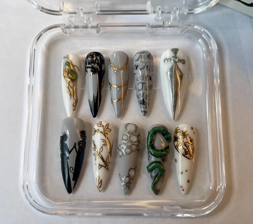 A set of ten press-on nails in a plastic, clear container. Three nails are white with gold accents, including a crown and a tree. Another nail has a white background and a painted silver dagger. The remaining nails are silver and feature different elements, such as the silhouette of a tree, the silhouette of two people, the word "jude" written multiple times and a green snake.