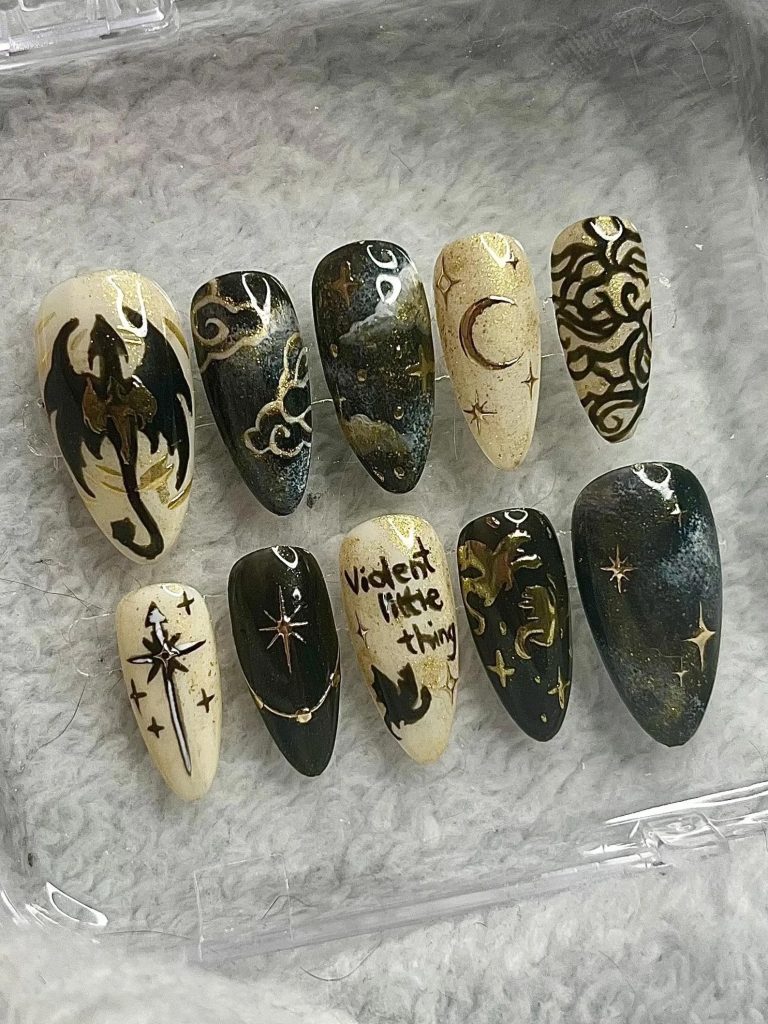 A Set of nails in a clear, plastic container. Five nails have a dark blue background with gold accents, such as stars and dragons. Five nails have a beige and gold background with black and gold accents, such as dragons, moons, and stars. One nail has the text "violent little thing" on it.