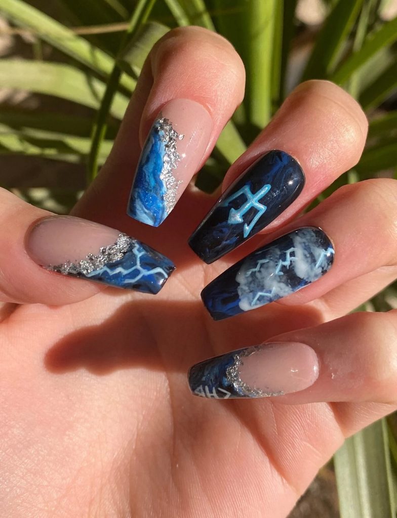 Nails against a leafy background. Two nails are a French tip style with different shades of blue and lighting on the tips and silver flakes separating the blue from a pale pink. Two nails are different shades of blue, one with a trident symbol and the other with clouds and lightning bolts. The last nail has a diagonal line of silver flakes separating blue marble polish and pale pink polish.