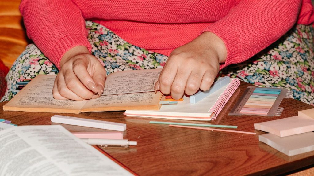 Image depicts a woman annotating a book with sticky tabs, surrounded by supplies like sticky notes, highlighters and a journal.