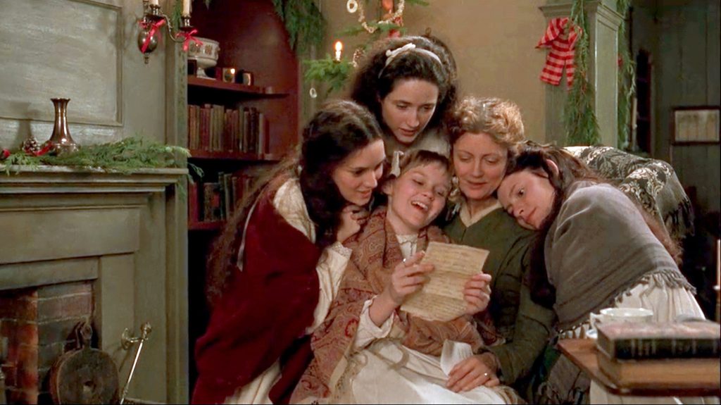 Movie still from Gillian Anderson's 1994 adaptation of 'Little Women'. Image depicting four young girls gathered around their mother as she sits in an arm chair. The smallest girl is reading from a letter. Christmas decorations adorn the cosy living room behind them.