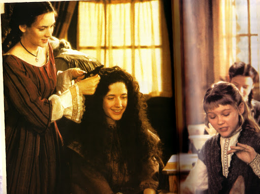 Movie still from Gillian Anderson's 1994 adaptation of 'Little Women'. Image depicting four young girls chatting together in a bedroom. One girl stands with curling tongs, curling her sisters hair. The other two girls are occupied with their own tasks.