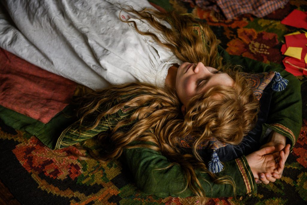 Movie still from Greta Gerwig's 2019 adaptation of 'Little Women'. Image depicting an auburn-curly-haired girl lying on a colourful patterned rug with her arms behind her head as she stares at the ceiling.
