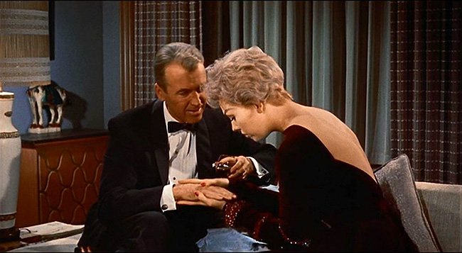 A still from the movie Bell, Book and Candle. A man with pale skin, gray hair, and a black tuxedo sits on a gray couch holding an alcoholic beverage. A woman with pale skin, short blonde hair, and a velvet burgundy evening dress sits beside him and leans down as she holds his hand with both of hers. The room around them includes green and brown curtains, a brown carved dresser with an animal statuette on top, and a white and gold lamp.