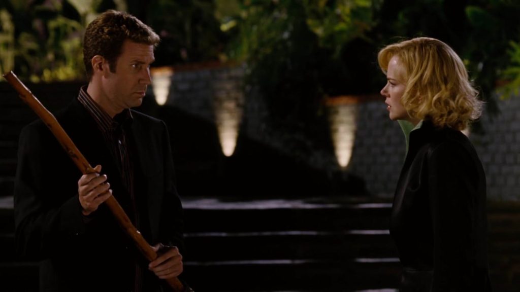 A still from the 2005 movie Bewitched. On the left, a man with pale skin, brown hair, and a black suit holds a broomstick and looks confused. Looking back at him from the right is a woman with blonde hair, pale skin, and a black coat trimmed in green. Behind them is a staircase with a pale brick wall, uplighting, and green plants.
