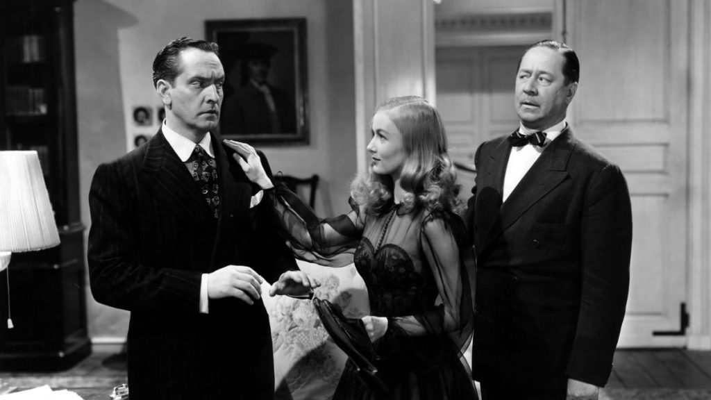 A black-and-white still from the movie I Married a Witch. A smiling woman with pale skin wearing a black dress with sheer sleeves places her hand on the shoulder of a concerned pale skinned man wearing a dark suit. Behind the woman is another man with pale skin and a dark suit looking on suspiciously.