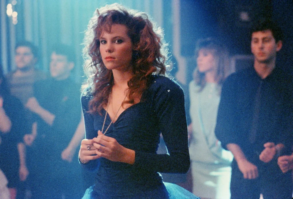 A still from the movie Teen Witch. A young girl with brown curly hair wears a dark blue prom dress and clutches the pendant on her necklace. She stands in a light blue spotlight. Behind her are other teens.