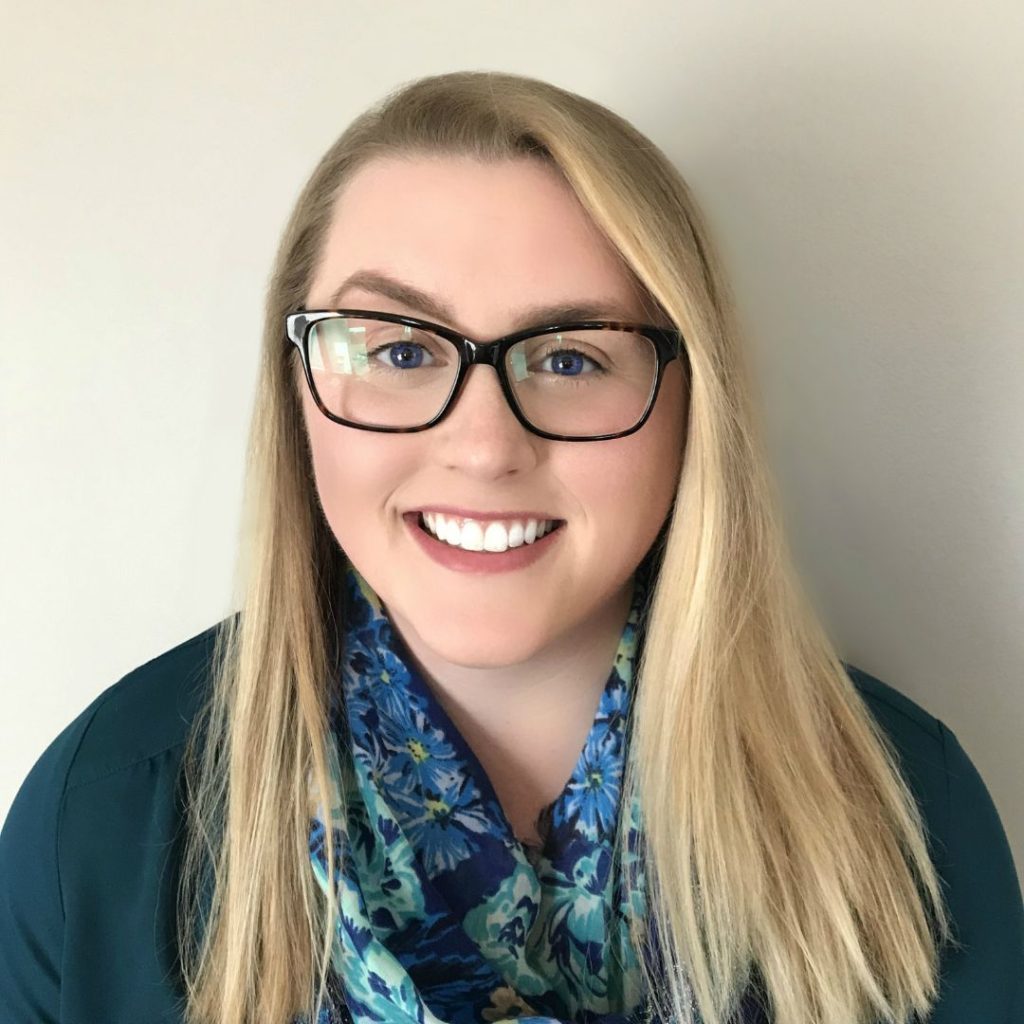 Close-up of Ali Novak, a fair-skinned blonde woman wearing black glasses, a floral blouse, and a green cardigan. She is smiling at the camera, and the background is beige.