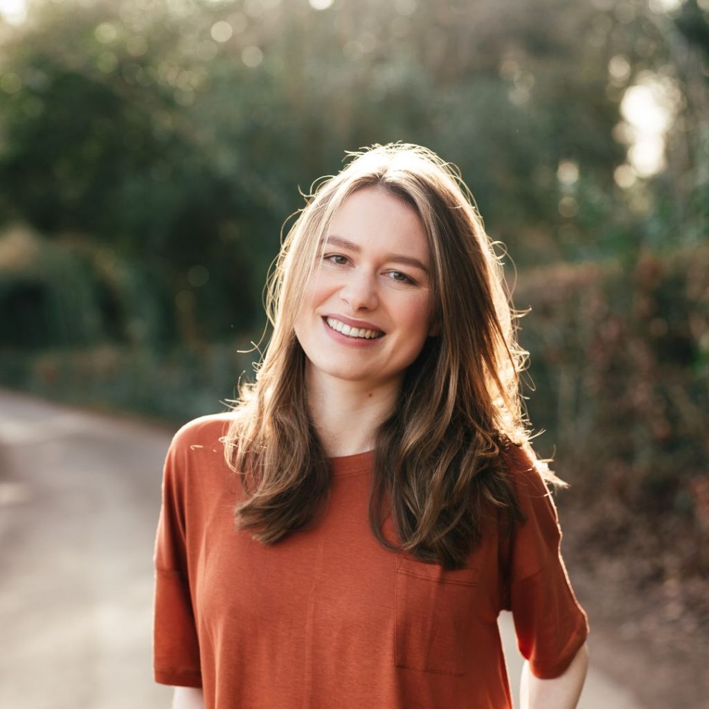 Close-up of Beth O'Leary, a fair-skinned woman with light brown hair wearing an orange shirt. She is smiling at the camera. The background is out of focus but depicts green trees and a path.