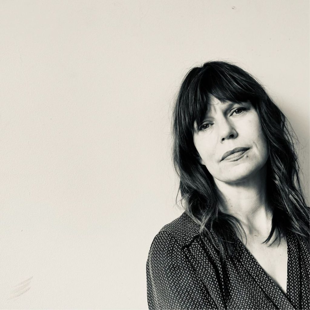 E. Lockhart, a woman with dark hair and bangs wearing a black and white blouse, staring at the camera. She is on the right side of the screen. The background is beige, and there is a beige filter over the whole image.