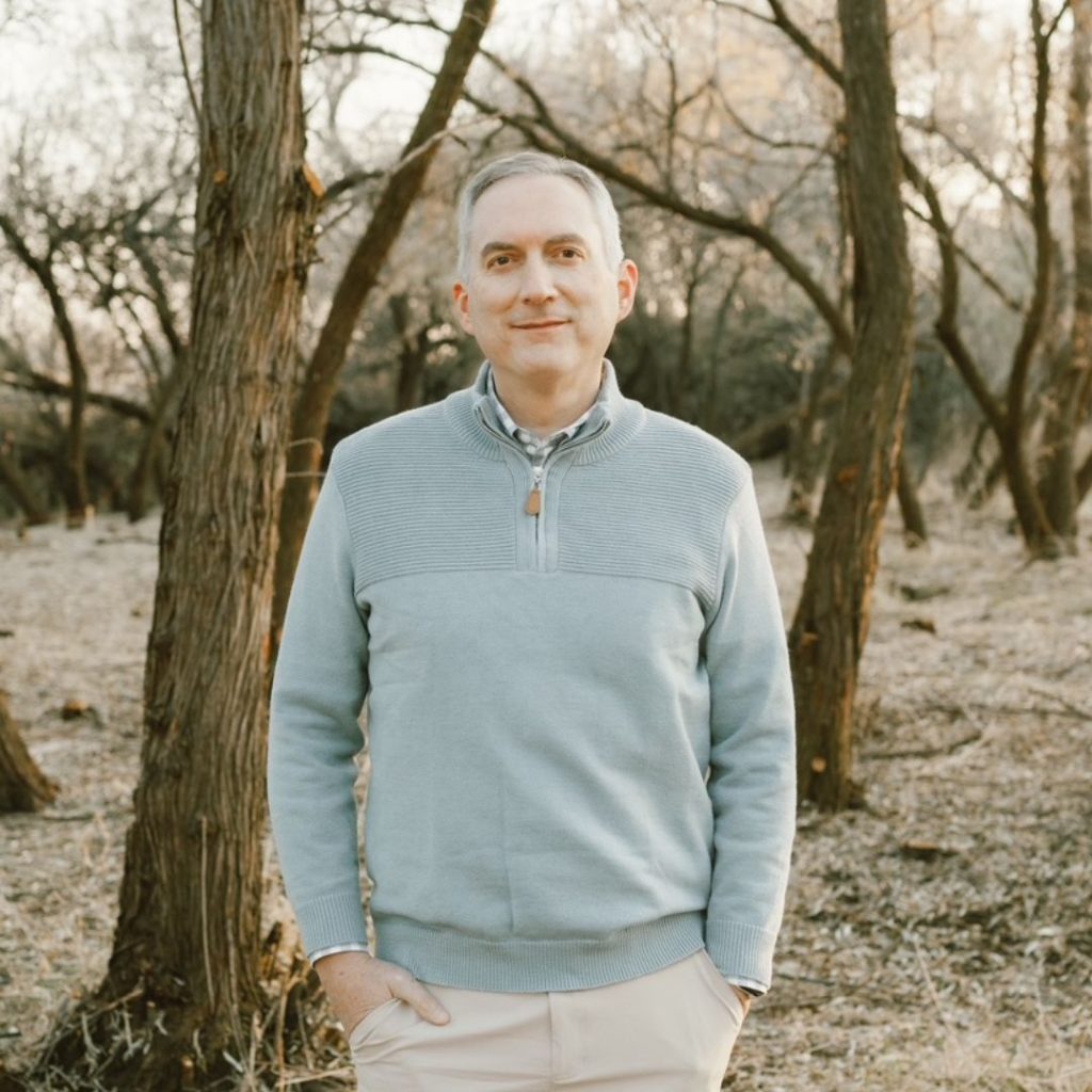 James Dashner, a fair-skinned man with short grey hair wearing a blue sweater and beige pants, smiling at the camera. The background consists of trees without leaves.