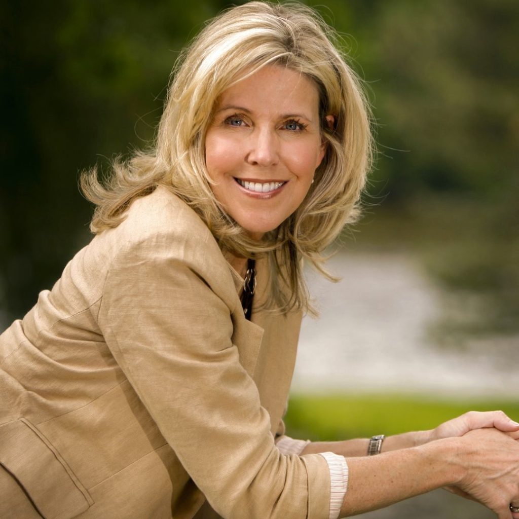 Lori Nelson Spielman, a fair-skinned woman with blonde hair wearing a beige blazer and beige pants, smiling at the camera with her head turned as she leans on something not shown. The background is out of focus, but is made up of different shades of green and grey.