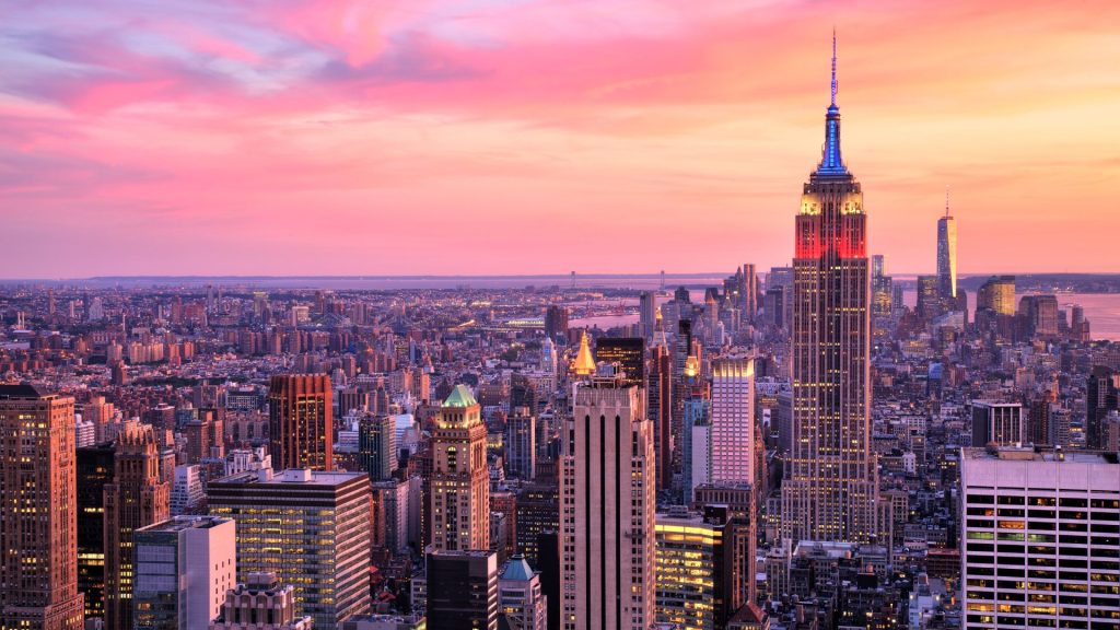 New York City skyline featuring the Empire State Building lit up in shades of blue, white, and red beneath a pink, dusky sky.