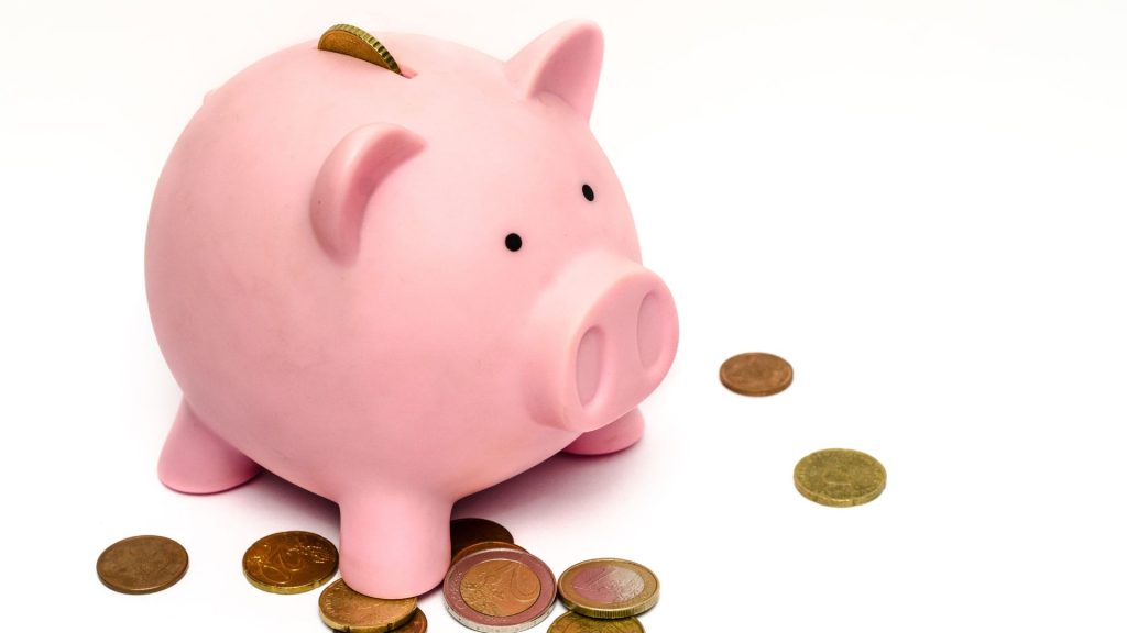A pink piggy bank with a coin sticking out of it stands facing to the right on a white background, surrounded by other coins.