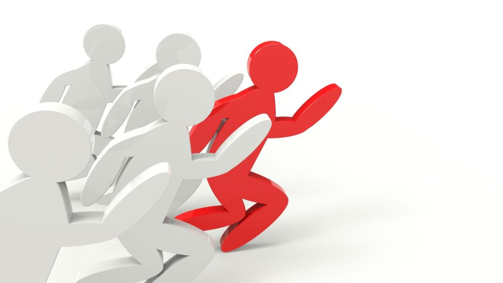 A group of five wooden figures, four white and one red, race on a white background. The red figure edges ahead of the rest of the pack.