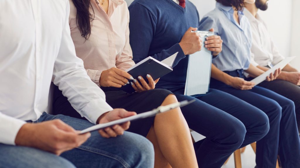 Five people in business attire sit in a row, each holding clipboards or a notebook, as they wait for a job interview.