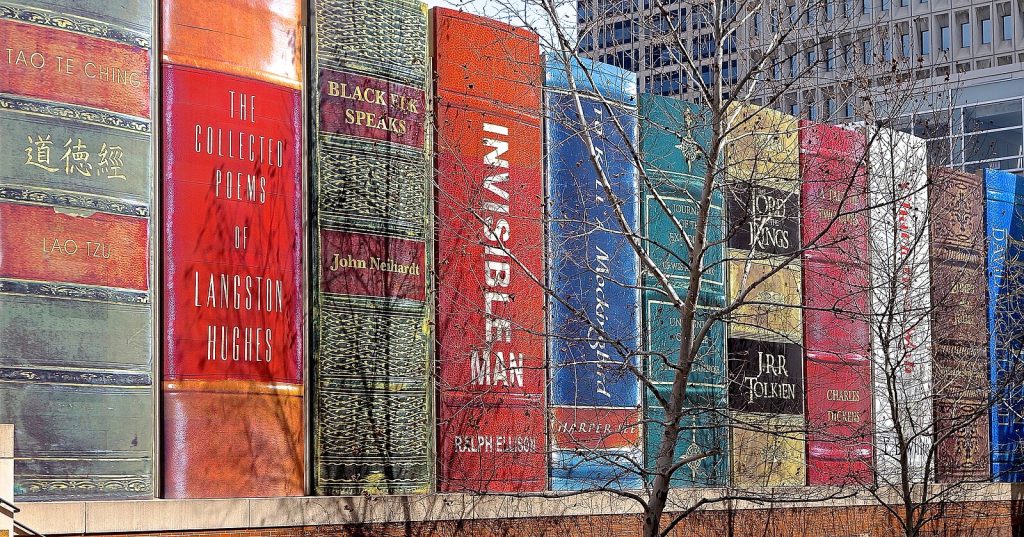 a picture of the Kansas City library parking garage decorated to look like a giant bookshelf, with books like Invisible man and To Kill A Mockingbird, slightly obscured by trees