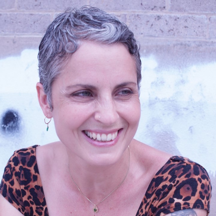 Anastasia Higginbotham, a smiling light-skinned woman with short gray hair and blue eyes, wearing a leopard-print short-sleeved shirt and sitting in front of a gray brick wall outdoors.