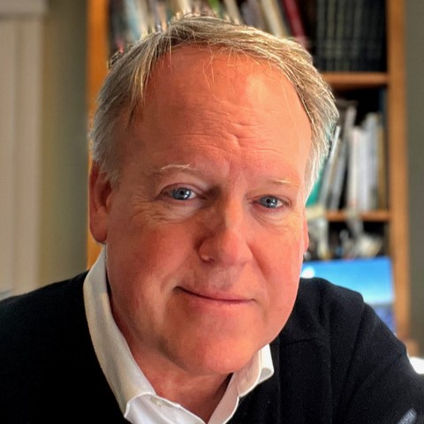 Brian Lies, a smiling light-skinned man with short silver hair and blue eyes, wearing a white collared shirt under a black sweater, posing in an office with bookshelves in the background.