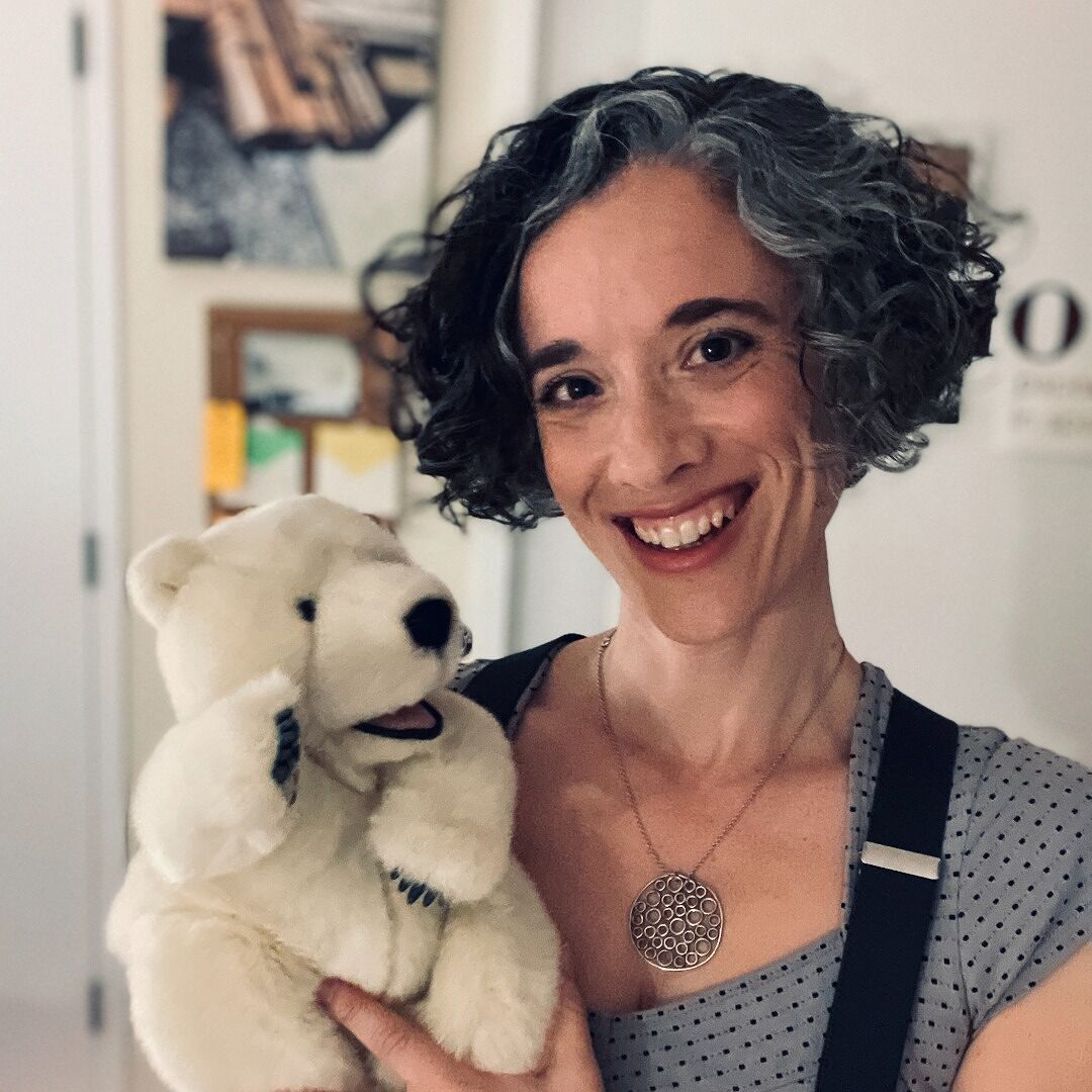 Caron Levis, a smiling light-skinned woman with short, curly salt-and-pepper hair and brown eyes, wearing a gray and black polka-dot shirt and black suspenders and holding a polar bear puppet while posing in an indoor studio area.