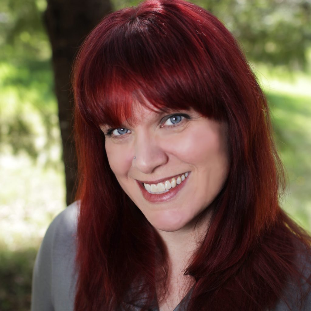 Lisa Mantchev, a smiling light-skinned woman with long, straight red hair and blue eyes, wearing a gray blouse and posing in a sunny, wooded outdoor area.