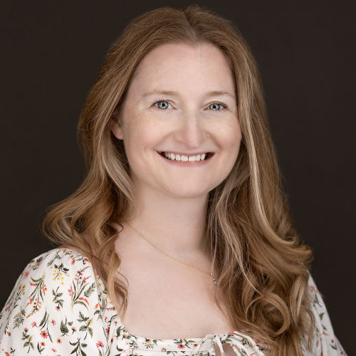 Headshot featuring Historical author Emily Bain Murphy. She appears against a darkened background, with a warm smile. Her light hair is styled in soft waves, framing her face naturally.