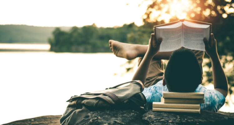 A tan-skinned child with short black hair reads a book while reclining on a lakeside rock, using several more books as a pillow. A knapsack lies nearby.