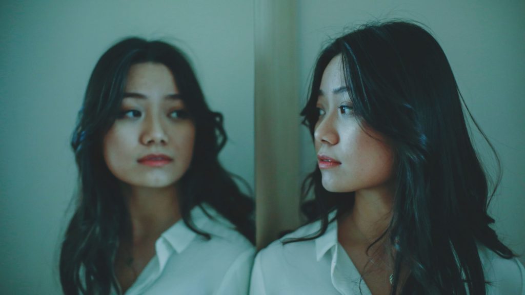 A young woman with light skin and long, black hair, wearing a white shirt, looks at herself in a mirror; the walls are grey and dimly lit.