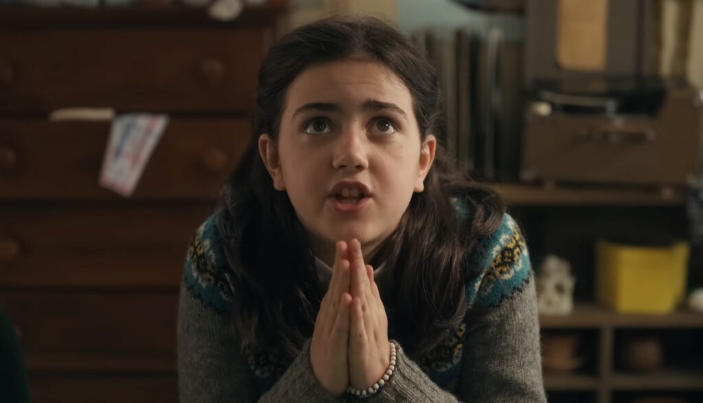 A still from the movie Are You There God? It's Me, Margaret. A young girl with dark brown hair and a gray sweater kneels in her bedroom and clasps her hands in prayer.