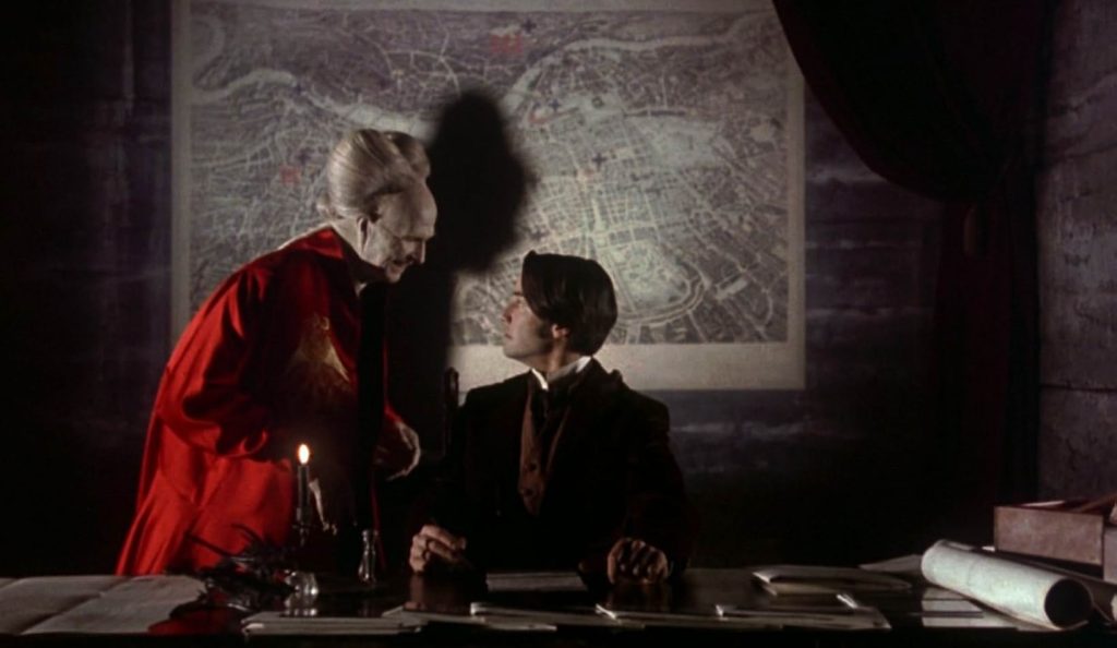 A still from the movie Bram Stoker's Dracula. A young man in a black suit and maroon vest sits at a desk and turns to face an old creature with white hair and skin wearing a bright red robe. On the wall behind them is an old fashioned metropolitan map.