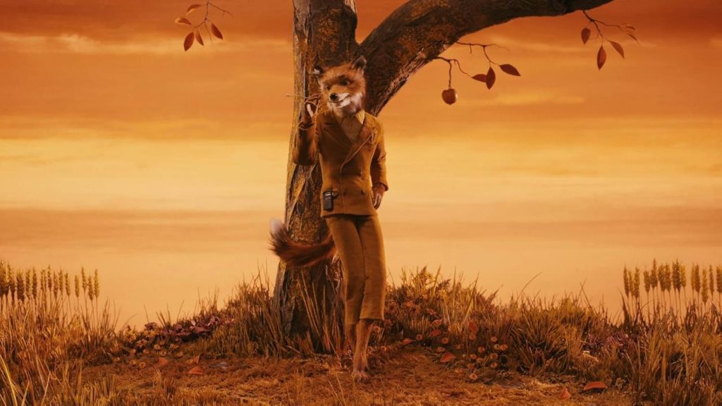 A still from the movie Fantastic Mr. Fox. A fox standing upright and wearing a brown suit with a walkman clipped to it leans against a tree with scattered fall leaves and chews on a piece of grass. Behind him is an orange and yellow sky.