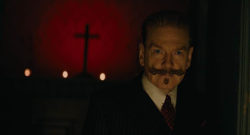 A still from the movie A Haunting in Venice. A man in a sharply tailor suit and manicured mustache stands in shadow and looks intense. Behind him is a cross backlit by red.