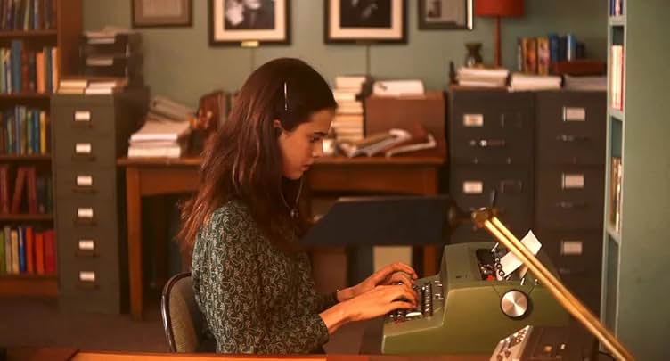 A still from the movie My Salinger Year. A young woman with long brown hair and a green blouse sits at a desk in a cluttered office and types on an olive green typewriter.