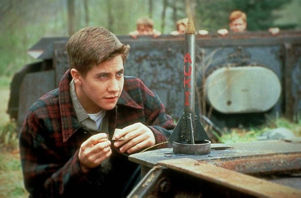 A still from the movie October Sky. A young man in a red and green flannel kneels in front of a small rocket and prepares to light its fuse. Crouched behind a metal structure are three other boys.
