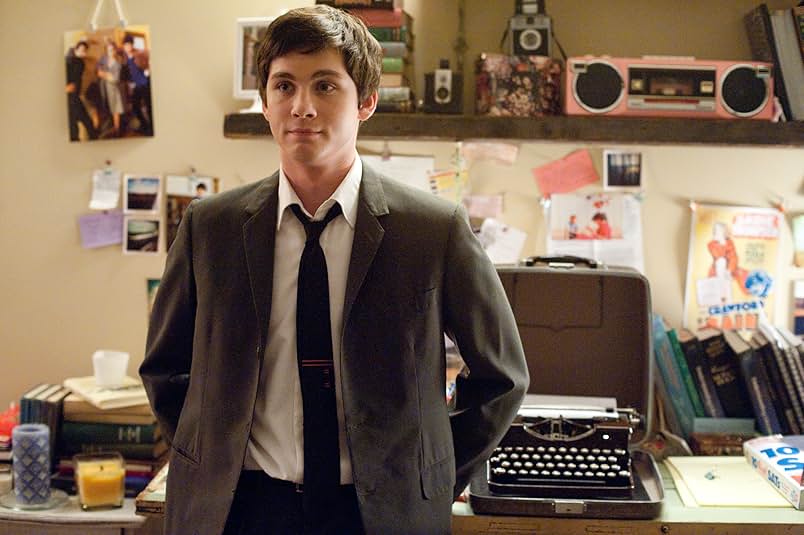 A still from the movie The Perks of Being a Wallflower. A teenage boy in a gray suit stands awkwardly in a colorful bedroom. Behind him is a typewriter, a boombox, books, and various drawings and photos pinned to the wall.