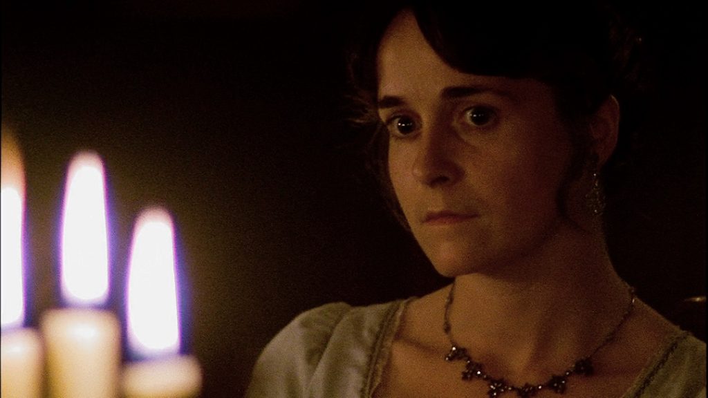 A still from the movie Persuasion. A woman with dark hair in Regency clothing sits in a dark room and faces a candelabra.