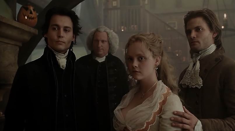 A still from the movie Sleepy Hollow. Three men and one woman in 18th century clothing stand in a large room lit by candles. On the fireplace mantle behind them is a carved jack-o-lantern.