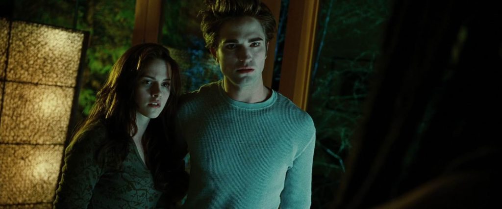 A still from the movie Twilight. A young woman in a green shirt stands next to a young man in a pale shirt who puts his arm around her protectively. Behind them are windows looking out onto forest scenery.