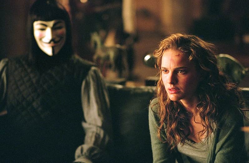 A still from the movie V for Vendetta. A man in a black puffed vest, gray blouse, Guy Fawkes mask, and brown wig sits on a couch and looks at a woman with golden curly hair wearing a green shirt.