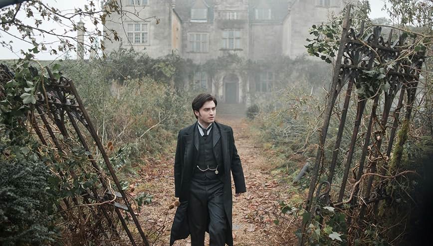 A still from the movie The Woman in Black. A man in a old fashioned black suit stands at the broken gate to a dilapidated estate. Behind him, the grounds are overgrown and covered in weeds and brambles.
