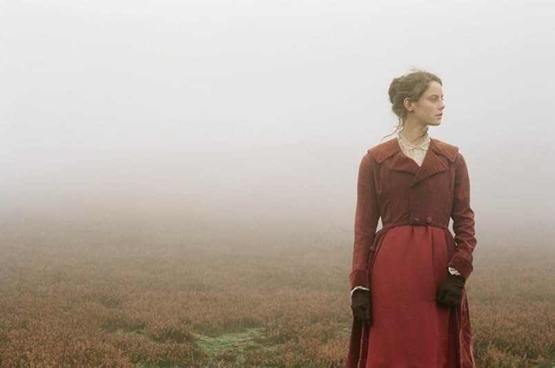 A still from the 2011 movie Wuthering Heights. A woman in a red and burgundy 19th century dress stands in a barren field surrounded by fog.
