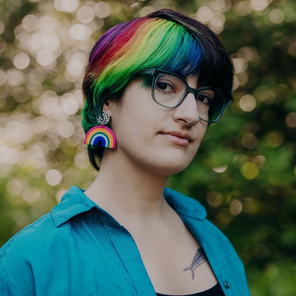Mar Romasco-Moore, a light-skinned individual with short dark hair dyed with rainbow stripes, a black shirt under a blue button-down, large teal glasses, and a rainbow earring, looks left at the viewer with their body turned three-quarters to the viewer's right. Trees can be seen out of focus in the background.