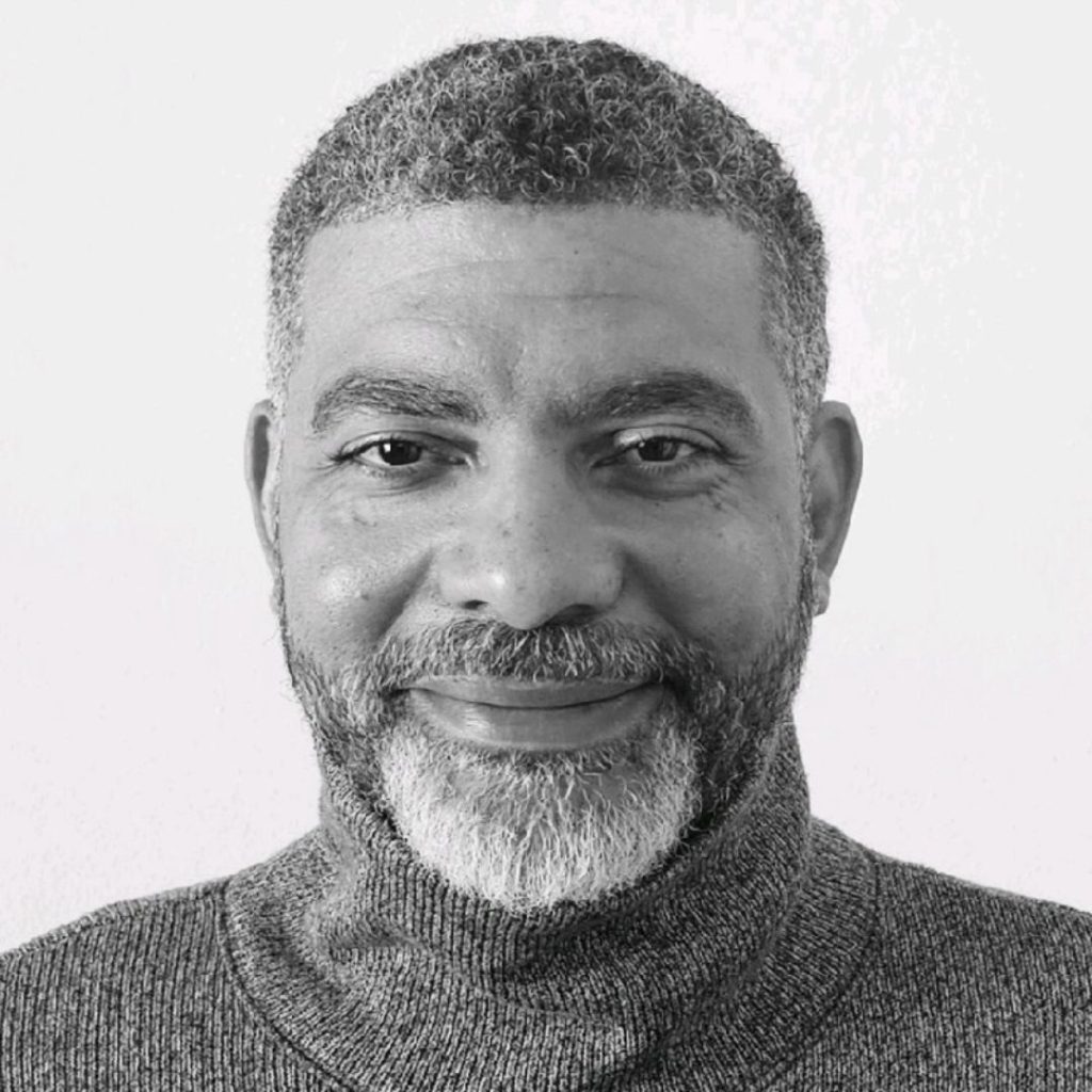 A black and white image of Thomas Jason Anderson wearing a turtleneck sweater and smiling slightly. He has short gray hair and short gray facial hair.