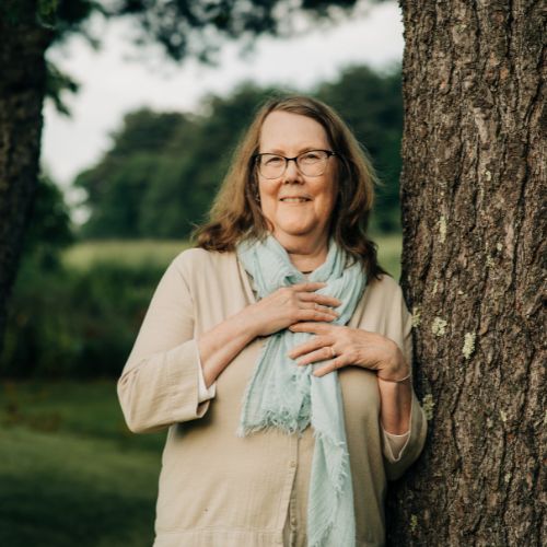 Headshot of Jeannine Atkins, a fair-skinned woman with brunette hair and black glasses, smiling at the camera. She is leaning against a tree, wearing a beige blouse with a blue scarf.