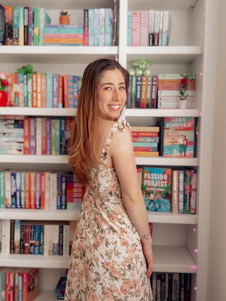Bookstagrammer Kailey turns her head over her shoulder with a beaming smile as she stands in front of her white bookshelf filled with colorful book covers. She is wearing a white dress with blush pink roses with green leaves.