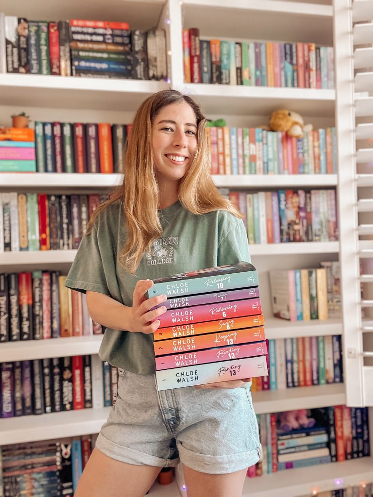 Kailey smiles in front of her vibrant bookshelf wearing a faded green shirt as she proudly displays Chloe Walsh's Boys of Tommen Series.