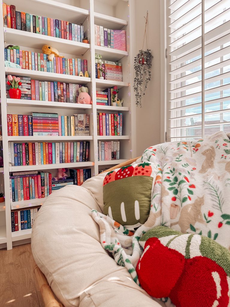 In a sun-soaked room, Kailey's white reading chair with strawberry and cherry pillows and a cozy blanket decorated with cats and plants sits in front of her colorful bookcase.