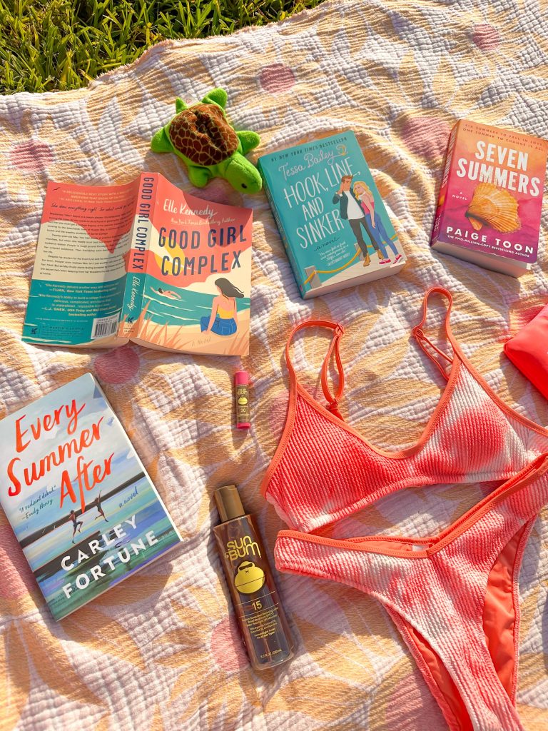 On a sun-soaked sunflower towel, a bottle of Sun Bum, a pink tie-dyed bikini, a small turtle stuffed animal, and four romance novels are laid out. From left to right is Every Summer After by Carley Fortune, Good Girl Complex by Ella Kennedy, Hook, Line, and Sinker by Tessa Bailey, and Seven Summers by Paige Toon.
